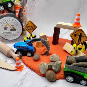 May include: A construction-themed sensory kit with toy trucks, cones, rocks, and wooden blocks. The kit includes a blue excavator, a green dump truck, and a sign that says "Detour Ahead". A rolling pin and a wooden knife are also included.
