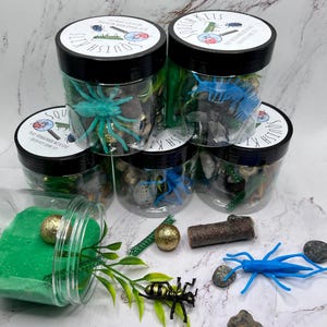 Op de afbeelding: Vijf doorzichtige potten gevuld met groene en bruine zintuiglijke items, waaronder plastic insecten, stenen en glitter. De potten hebben zwarte deksels met witte tekst die "Squish Kits" luidt.