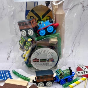 May include: A clear plastic bag filled with a train-themed play dough kit. The kit includes toy trains, wooden blocks, green pipe cleaners, and a butterfly decoration. The bag has the text "SQUISH KITS" and "PLAY DOUGH MADE WITH LOVE".