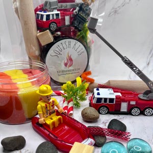 May include: A play dough kit with a firefighter theme. The kit includes a red fire truck toy, a firefighter figurine, a red inflatable boat, and colorful play dough. The kit also includes small rocks, wooden blocks, and decorative elements.