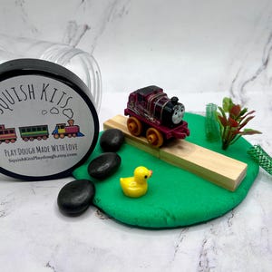May include: A play dough kit with a toy train, a yellow rubber duck, and black stones on a green surface. The kit includes a container with a black lid labeled "Squish Kits" and the text "Play Dough Made With Love."