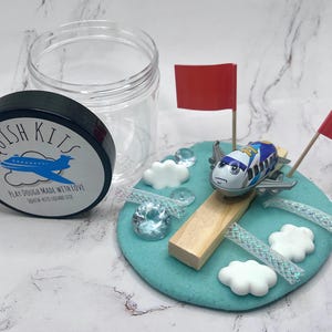 Puede incluir: Un tarro de plástico transparente con una tapa negra que dice "Squish Kits Play Dough Made With Love Squish-Kits Square Size" y un juguete de avión azul y blanco sobre una superficie de plastilina azul con nubes blancas, un palo de madera y una cuerda iridiscente.