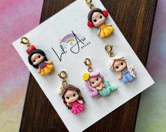 May include: Six colorful polymer clay charms featuring cartoon characters. The charms include Snow White, a mermaid, and other princesses. Each charm has a gold-colored clasp. The charms are displayed on a white card with the Lil' Ari Design logo.