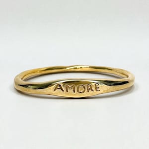Op de afbeelding: Een goudkleurige ring met een gepolijste, organische en licht onregelmatige band. Het voorste gedeelte van de ring is afgeplat en voorzien van het woord AMORE gegraveerd in hoofdletters. Het metalen oppervlak heeft een reflecterende afwerking.