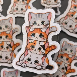 May include: A stack of four cat stickers. The top cat is gray with pink ears, the second is orange, the third is white with orange spots, and the bottom cat is white with orange spots. The stickers have a white border and are on a dark gray background.