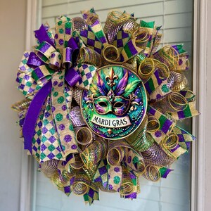 Puede incluir: Una corona de Mardi Gras con un medall&oacute;n central con un dise&ntilde;o de m&aacute;scara. La corona est&aacute; compuesta por cintas moradas, verdes y doradas con lunares y patrones de cuadros. El medall&oacute;n tiene las palabras "Mardi Gras".