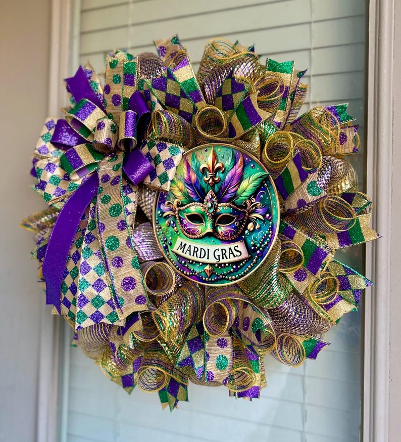 Puede incluir: Una corona de Mardi Gras con un medall&oacute;n central con un dise&ntilde;o de m&aacute;scara. La corona est&aacute; compuesta por cintas moradas, verdes y doradas con lunares y patrones de cuadros. El medall&oacute;n tiene las palabras "Mardi Gras".