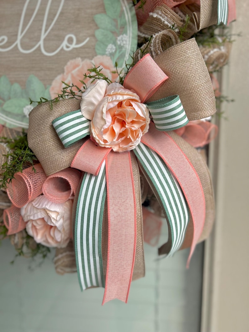 May include: A peach and green striped ribbon bow with a peach flower in the centre. The bow is tied around a burlap wreath with a sign that says "hello".