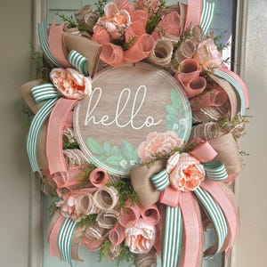 May include: A peach and green wreath with a wooden sign that says "hello" in white lettering. The wreath is decorated with burlap, peach-coloured ribbon, and faux flowers.