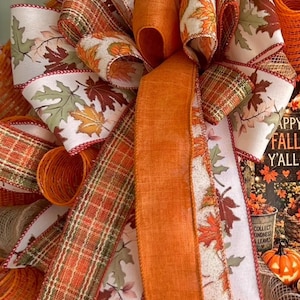 May include: A decorative wreath featuring a large central bow made of orange, plaid, and white ribbon with a printed autumn leaf pattern. The wreath includes orange mesh loops and a sign with the text HAPPY FALL Y'ALL and COLLECT KINDNESS LEAVES.