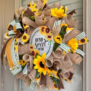 May include: A burlap wreath decorated with sunflowers, brown and green striped ribbon, and a round sign that reads "Home Sweet Home".