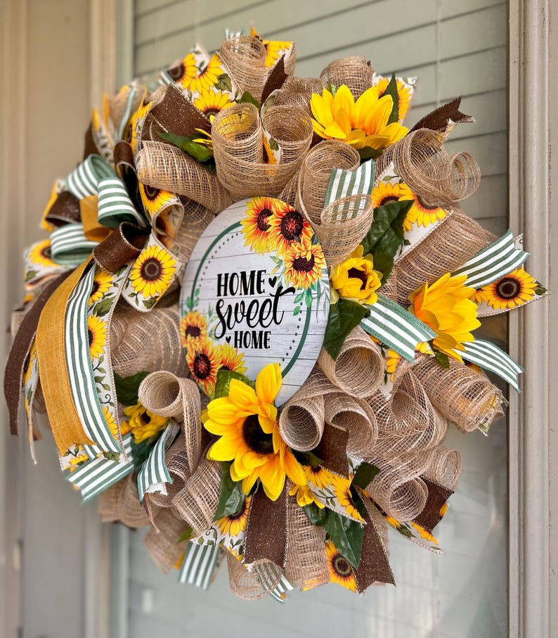 May include: A burlap wreath decorated with sunflowers, brown and green striped ribbon, and a round sign that reads "Home Sweet Home".