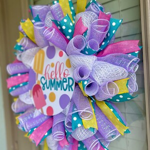 May include: A summer wreath with a purple, white, teal, and yellow mesh base. The wreath has a round sign in the center that says "hello summer" with a popsicle graphic.