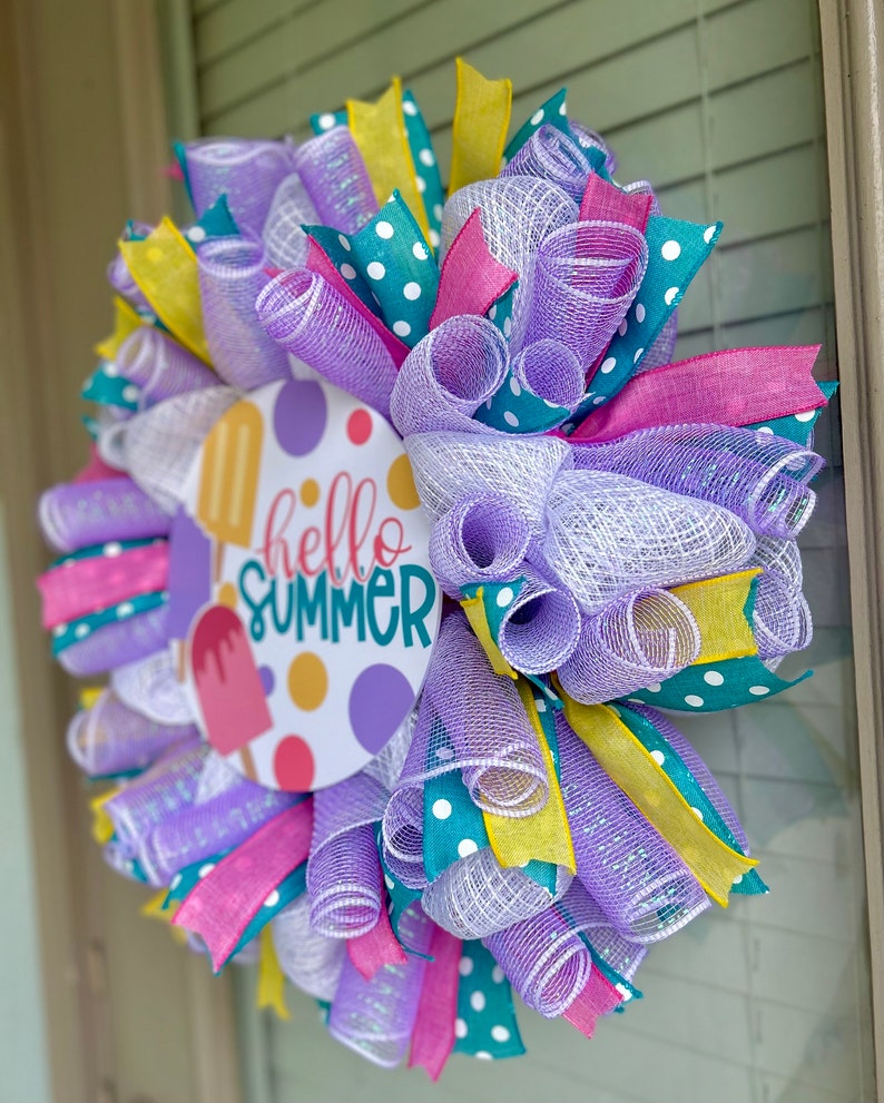 May include: A summer wreath with a purple, white, teal, and yellow mesh base. The wreath has a round sign in the center that says "hello summer" with a popsicle graphic.