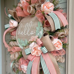 May include: A peach and green wreath with a wooden sign that says "hello". The wreath is made with burlap, mesh, and ribbon.