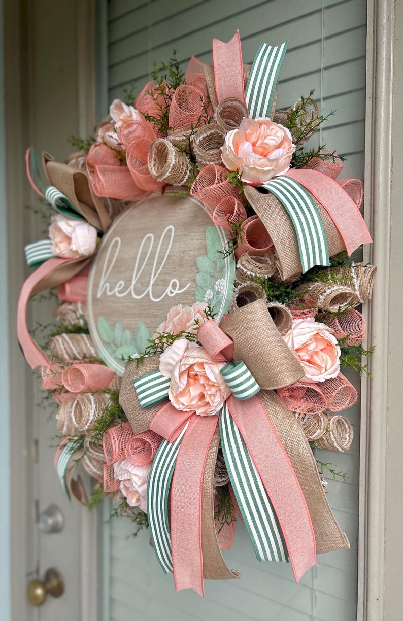 May include: A peach and green wreath with a wooden sign that says "hello". The wreath is made with burlap, mesh, and ribbon.