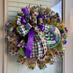 Puede incluir: Una festiva corona de Mardi Gras con cintas moradas, verdes y doradas. La corona presenta una m&aacute;scara decorativa con las palabras "Mardi Gras" y est&aacute; adornada con varias cintas y lazos estampados, creando un aspecto festivo.