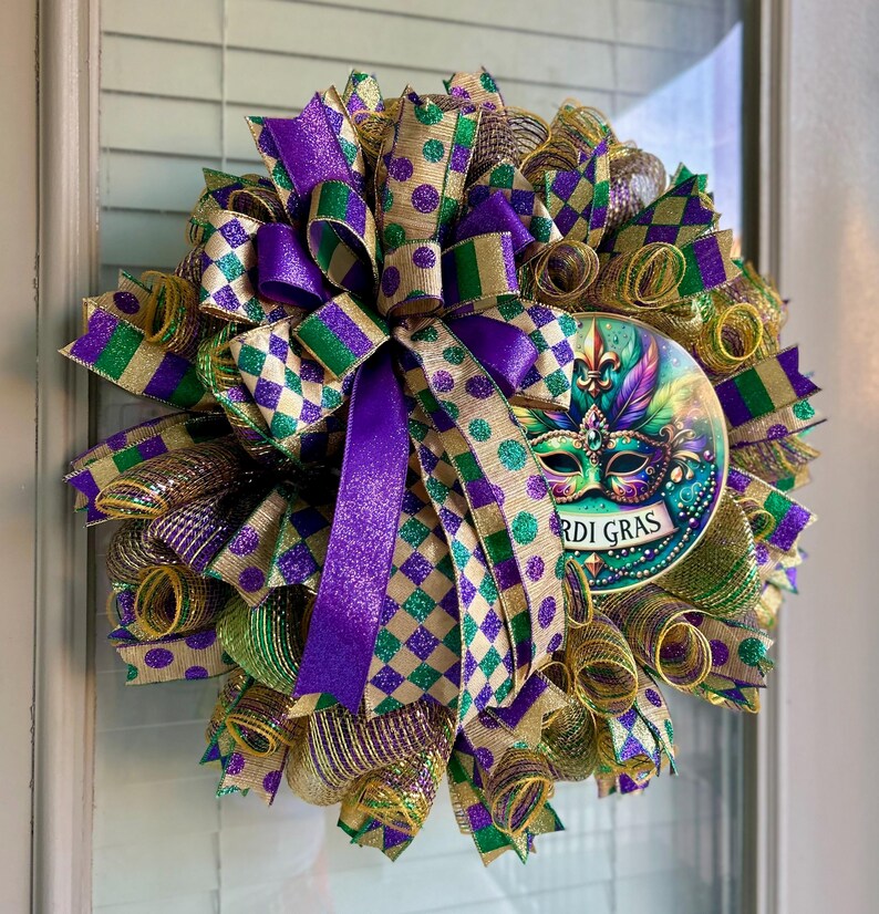 Puede incluir: Una festiva corona de Mardi Gras con cintas moradas, verdes y doradas. La corona presenta una m&aacute;scara decorativa con las palabras "Mardi Gras" y est&aacute; adornada con varias cintas y lazos estampados, creando un aspecto festivo.