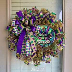 Puede incluir: Una corona de Mardi Gras con un dise&ntilde;o de m&aacute;scara central. La corona presenta una vibrante mezcla de cintas y mallas moradas, verdes y doradas. La m&aacute;scara est&aacute; detallada con plumas y las palabras "Mardi Gras".