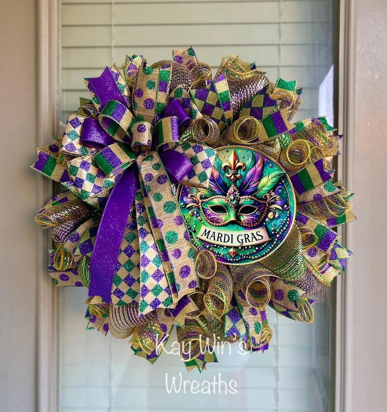 Puede incluir: Una corona de Mardi Gras con un dise&ntilde;o de m&aacute;scara central. La corona presenta una vibrante mezcla de cintas y mallas moradas, verdes y doradas. La m&aacute;scara est&aacute; detallada con plumas y las palabras "Mardi Gras".