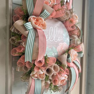 May include: A peach and green wreath with a burlap base and a sign that says "hello". The wreath is decorated with peach-coloured flowers and green and white striped ribbon.
