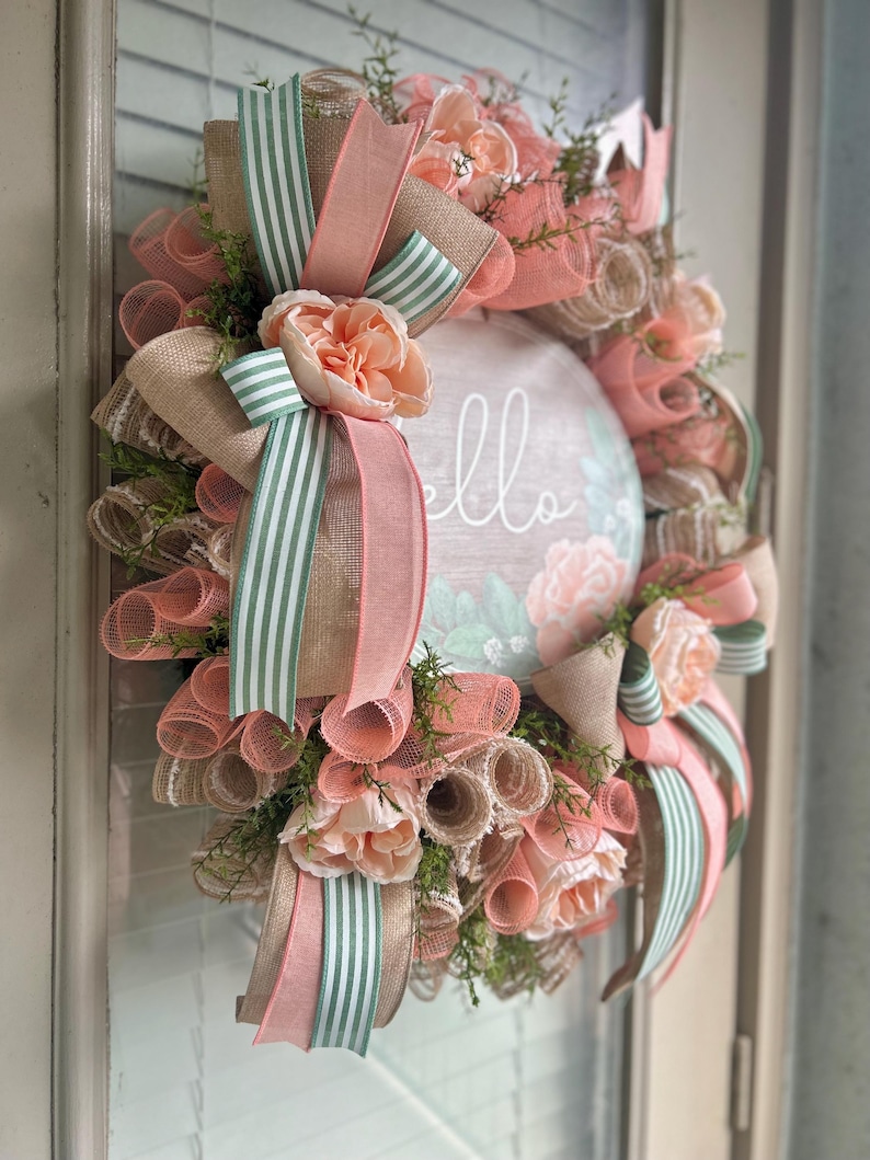 May include: A peach and green wreath with a burlap base and a sign that says "hello". The wreath is decorated with peach-coloured flowers and green and white striped ribbon.