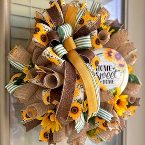 May include: A sunflower wreath with a brown, yellow, and green ribbon bow. The wreath has a wooden sign that says "Home Sweet Home".