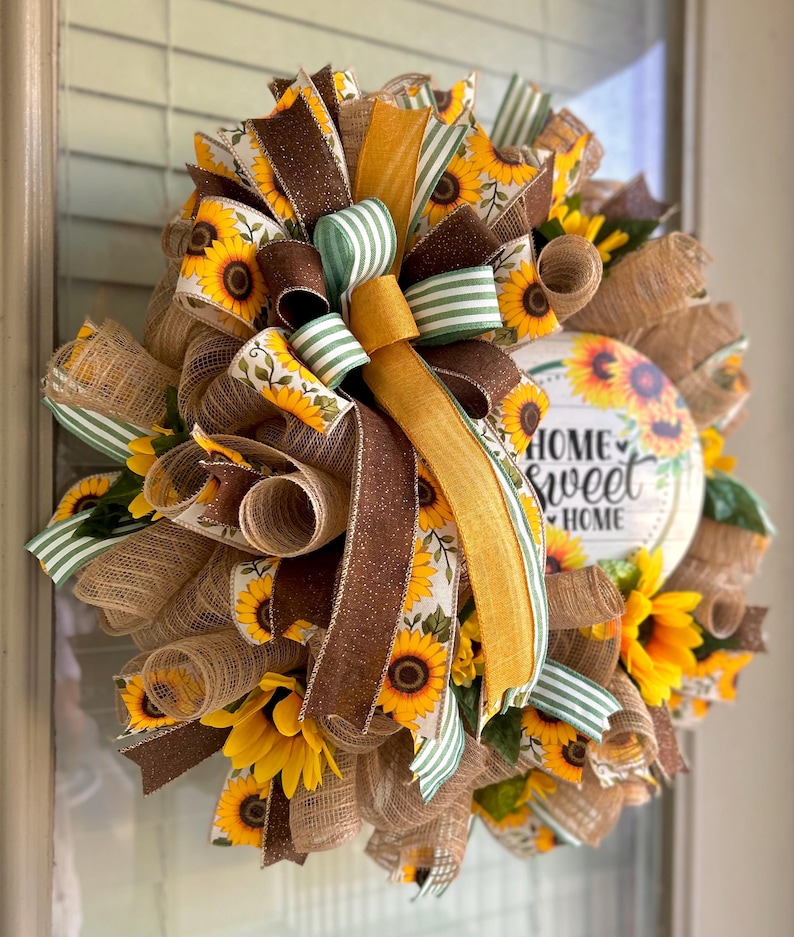 May include: A sunflower wreath with a brown, yellow, and green ribbon bow. The wreath has a wooden sign that says "Home Sweet Home".