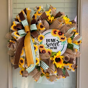 May include: A sunflower wreath with a brown and green ribbon bow. The wreath has a round sign in the center that says "Home Sweet Home" with a small heart.