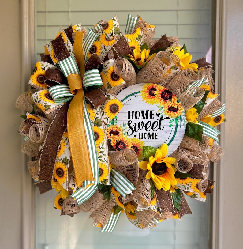 May include: A sunflower wreath with a brown and green ribbon bow. The wreath has a round sign in the center that says "Home Sweet Home" with a small heart.