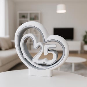 Könnte beinhalten: Ein weiß-graues herzförmiges Deko-Objekt mit der Zahl 25. Das Objekt steht auf einem weißen Sockel und befindet sich auf einem weißen Tisch. Der Hintergrund ist ein verschwommenes Wohnzimmer.