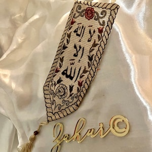 May include: A beige fabric wall hanging with a tassel. The fabric has a brown rope border and an embroidered design with the Arabic words "Allah" and "La ilaha illa Allah" in red thread. The wall hanging is decorated with small red flowers. The word "Jahuri" is written in gold letters on a white surface below the wall hanging.