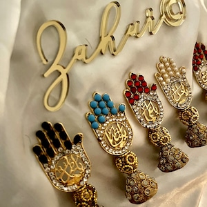 Könnte beinhalten: Fünf goldene Hamsa-Hand-Anhänger mit verschiedenen farbigen Steinen und Strasssteinen. Jeder Anhänger hat arabische Kalligraphie in der Mitte. Die Anhänger befinden sich auf einem weißen Hintergrund.