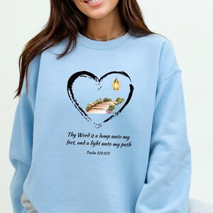 May include: Light blue sweatshirt with a heart-shaped graphic featuring a path with a lantern and the text "Thy Word is a lamp unto my feet, and a light unto my path Psalm 119:105".