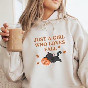 May include: Cream-colored hoodie with the text "JUST A GIRL WHO LOVES FALL" in orange. The graphic features a black cat hugging a pumpkin. The hoodie has a drawstring hood and a front pocket.