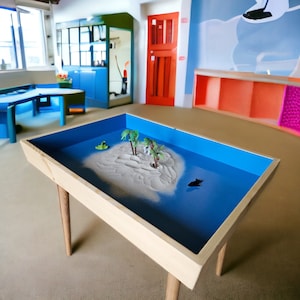 May include: A wooden sandbox with a blue interior and a small sand island with palm trees. The sandbox is on a light brown floor in a room with colorful furniture.