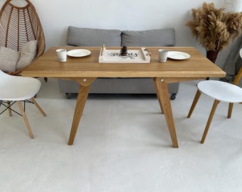 Puede incluir: Una mesa de comedor de madera clara con patas anguladas. La mesa está puesta con platos blancos, tazas y una bandeja decorativa. Dos sillas blancas y una silla de mimbre están colocadas alrededor de la mesa. La mesa mide unos 152 cm de largo.