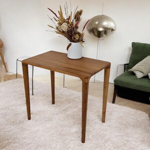 May include: A rectangular wooden table with a natural finish. The table has four tapered legs and rounded corners. A white vase with dried flowers sits on top. The table is in a room with a green sofa and a fluffy rug.