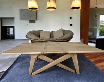 Puede incluir: Una mesa de comedor de madera clara con una base única de patas cruzadas. La mesa rectangular parece extensible. Está colocada sobre una alfombra azul en una habitación bien iluminada, con un sofá redondo en el fondo. La mesa mide unos 150 cm de largo.