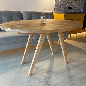 Puede incluir: Una mesa ovalada de madera clara con un diseño simple y moderno. La mesa tiene cuatro patas que forman una forma triangular. La mesa está sobre un suelo de madera clara.