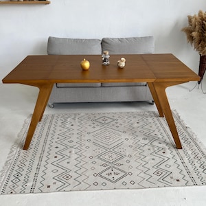 May include: A wooden dining table with a light brown finish and a geometric pattern rug underneath. The table has a unique design with angled legs and a rectangular top.
