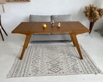 Puede incluir: Una mesa de comedor de madera con un acabado marrón claro y una alfombra con un patrón geométrico debajo. La mesa tiene un diseño único con patas anguladas y una tapa rectangular.