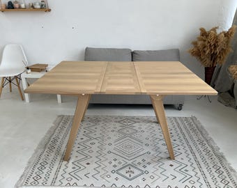 Puede incluir: Una mesa de comedor de madera clara con una tapa rectangular y tres patas a cada lado. La mesa está colocada sobre una alfombra blanca con un patrón geométrico.