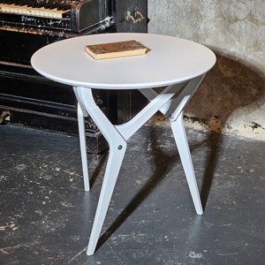 May include: A white round table with a white top and four legs. The table is made of wood and has a simple design. A book is sitting on the table.