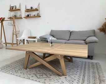 Puede incluir: Una mesa de centro de madera clara con una base en forma de X única. La mesa está hecha de madera natural y tiene un acabado marrón claro. La mesa está sobre una alfombra de patrones blancos y grises en un salón.