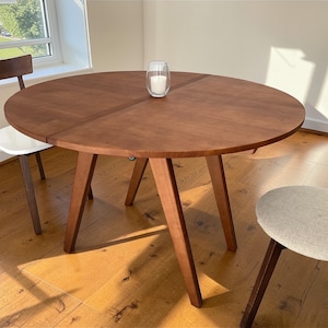 May include: A round wooden dining table with a warm brown finish, supported by three angled legs. Two chairs are visible, one with a white seat and the other with a light gray seat. A glass candle holder sits on the table.