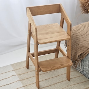 Könnte beinhalten: Ein hellfarbiger Hochstuhl aus Holz mit Fußstütze und verstellbarem Sitz. Der Stuhl hat eine Rückenlehne und Armlehnen. Der Stuhl ist aus Naturholz gefertigt und für ein Kind konzipiert.