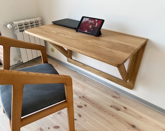 May include: A light wood wall-mounted desk with a laptop and tablet on top. The desk has a simple design with a triangular support structure. A wooden chair with a gray cushion sits in front of the desk.