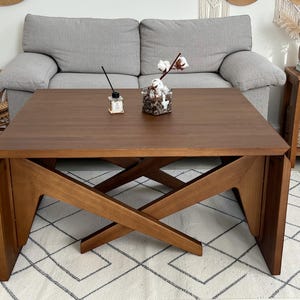 May include: A square, wooden coffee table with a dark brown finish. The table has an X-shaped base and is placed on a white rug with a geometric pattern. A gray sofa is in the background.