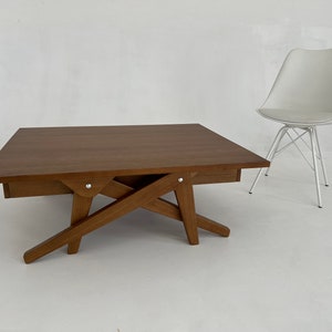 May include: A rectangular wooden coffee table with a dark brown finish and a unique scissor-style base featuring silver metal hardware, positioned next to a white molded plastic chair with a white metal wire base and a light gray seat cushion.